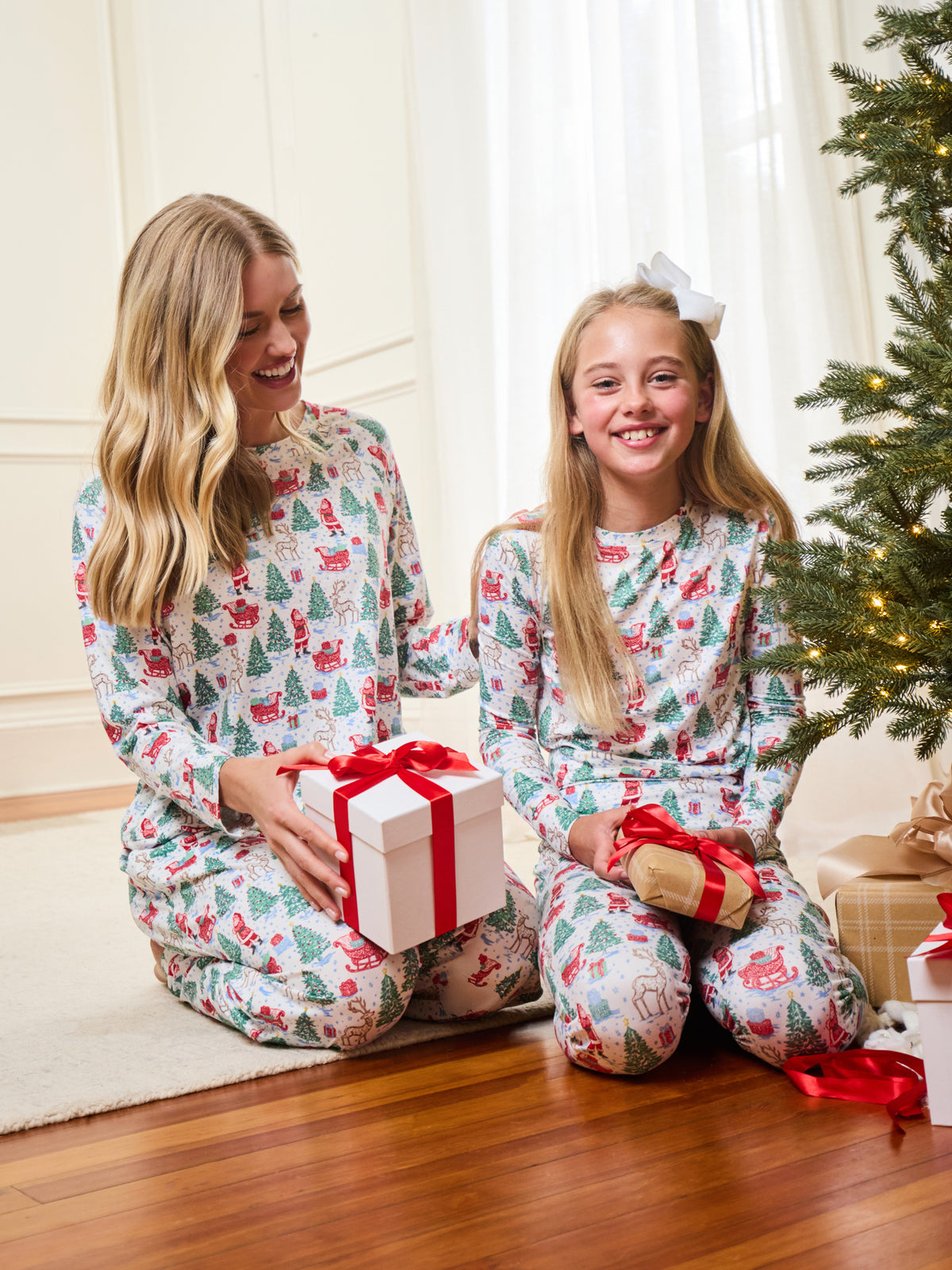 A woman and a young girl sitting on a wooden floor in a living room, both wearing matching long-sleeve pajamas with a festive Christmas tree and Santa Claus pattern. The girl holds a wrapped gift with a red ribbon, and a Christmas tree is visible in the background.