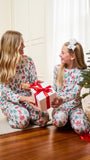 A woman and a young girl sitting on a wooden floor in a cozy living room, both wearing matching Christmas-themed pajamas with a Santa Claus and tree pattern. The girl is holding a white gift box with a red ribbon, with a Christmas tree and wrapped presents in the background.