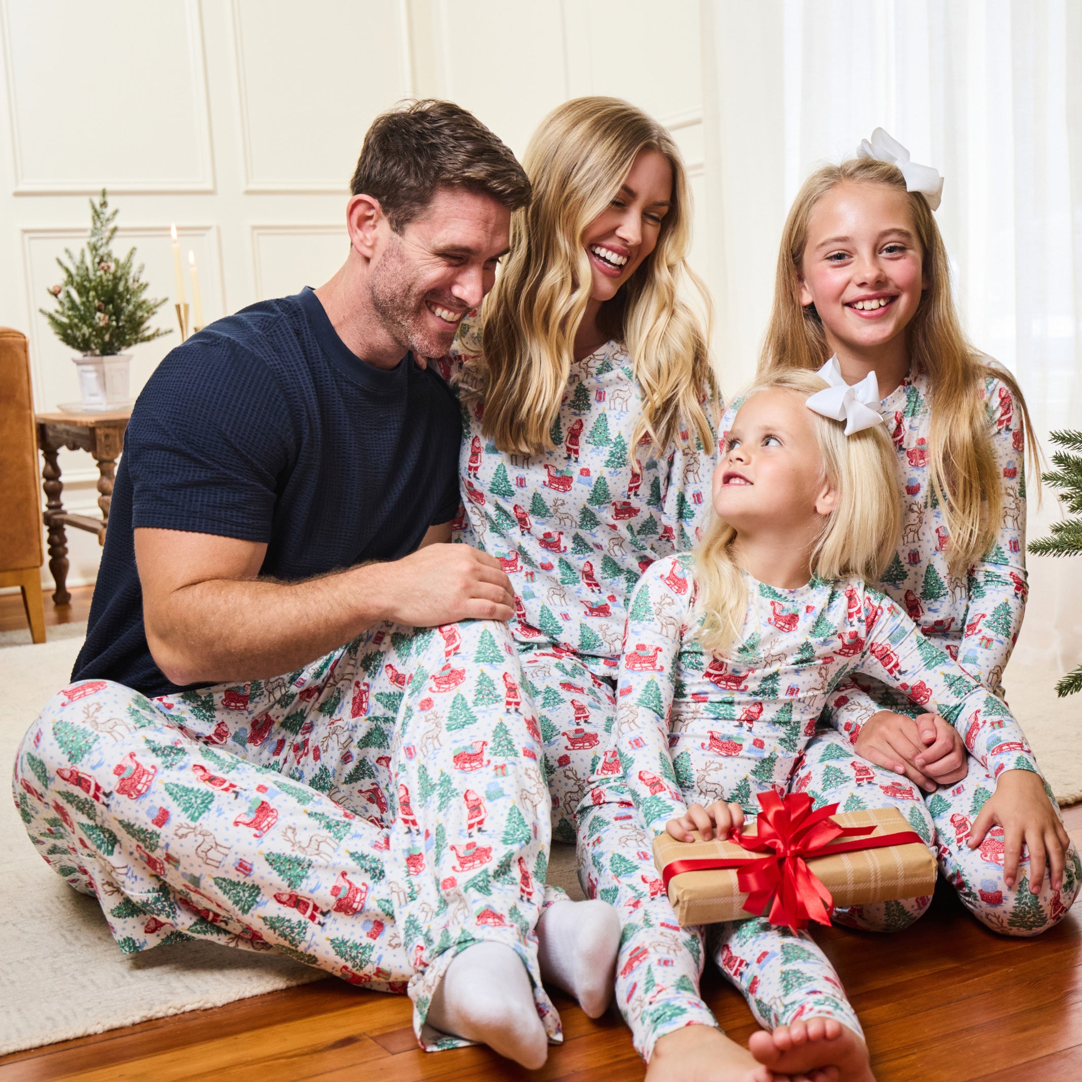 A family of four wearing matching Christmas-themed pajamas sits together on a wooden floor in a bright living room. The pajamas have a white base with a festive print of Santa Claus, reindeer, snow globes, and Christmas trees, with red accents.
