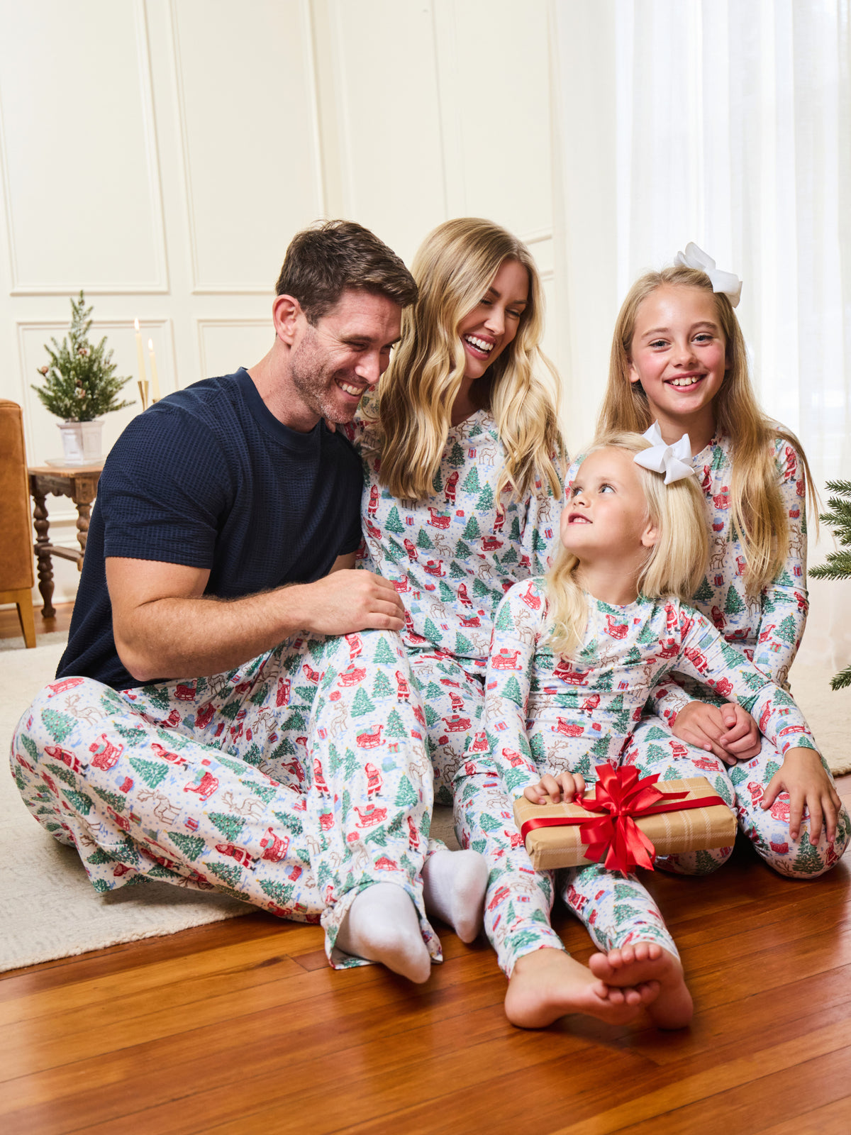 A family of four wearing matching Christmas-themed pajamas sits together on a wooden floor in a bright living room. The pajamas have a white base with a festive print of Santa Claus, reindeer, snow globes, and Christmas trees, with red accents.