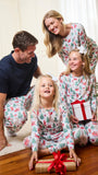 A family of four wearing matching Christmas-themed pajamas sits together on a wooden floor in a living room, smiling and celebrating around wrapped presents near a decorated Christmas tree.