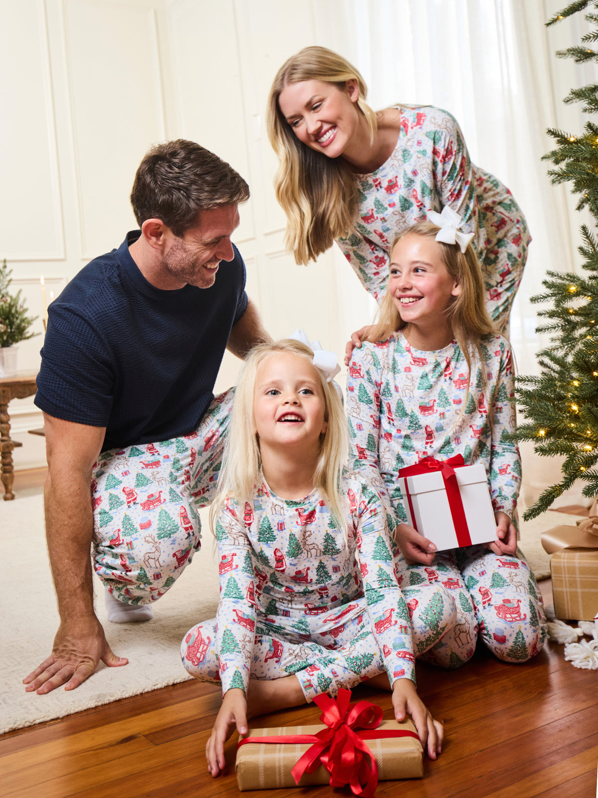 A family of four wearing matching Christmas-themed pajamas sits together on a wooden floor in a living room, smiling and celebrating around wrapped presents near a decorated Christmas tree.