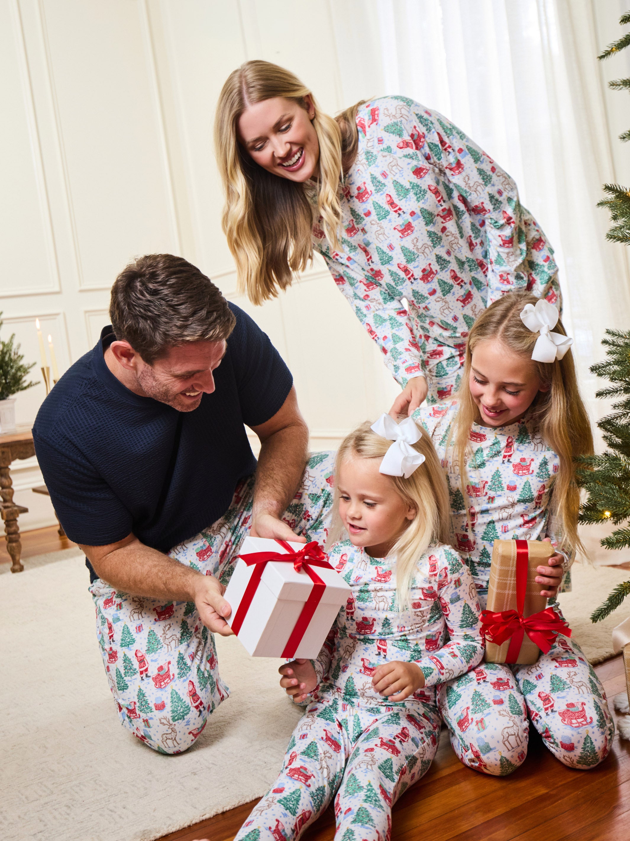 A family of four wearing matching Christmas-themed pajamas with a green pine tree and red berry pattern, gathered in a living room around a wrapped gift box. The mother and daughter on the right wear white bows in their hair, and the father kneels on the left.