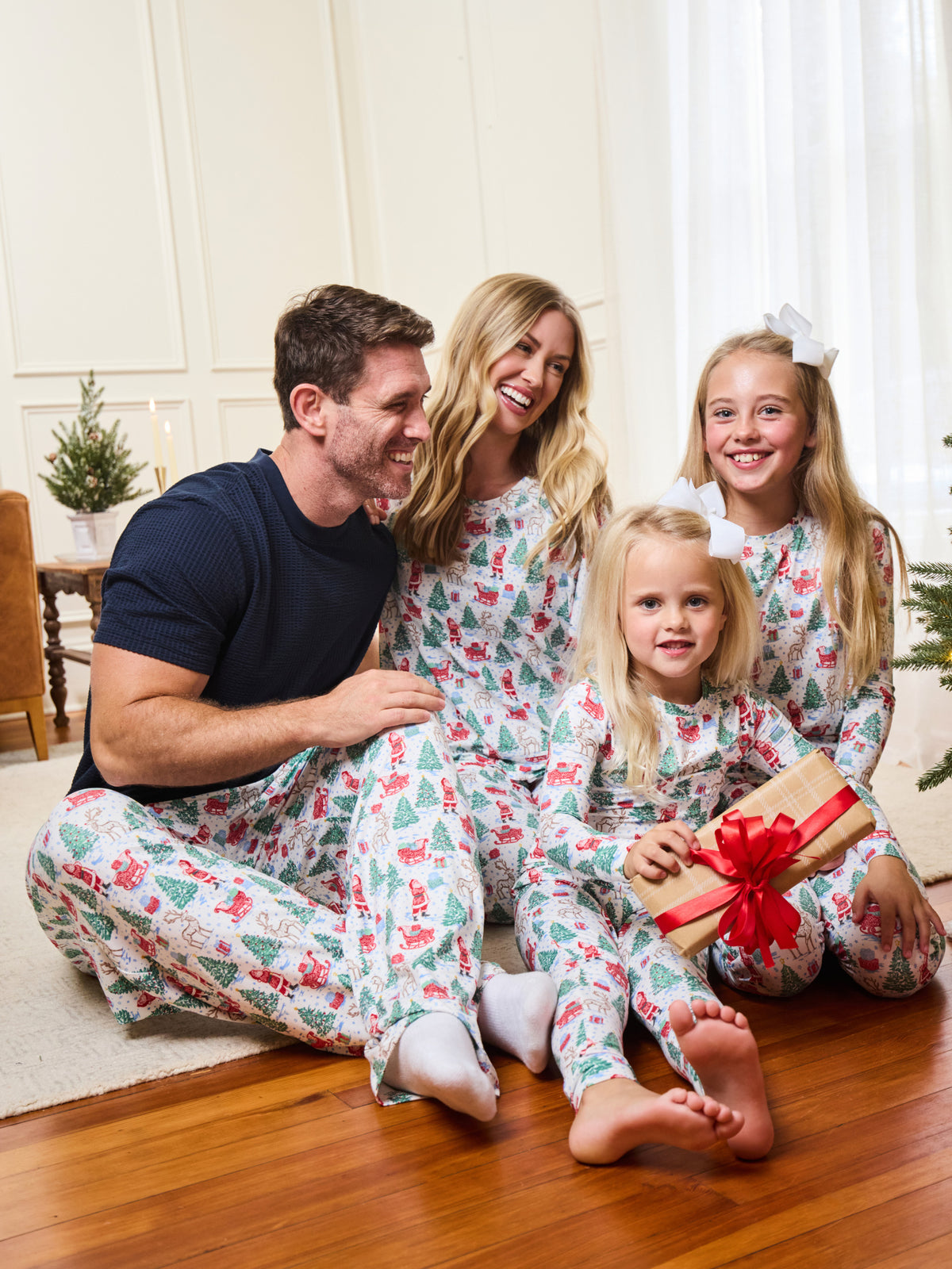 A family of four wearing matching holiday-themed pajamas sits together on a wooden floor in a bright living room. The pajamas have a white base with a festive red, green, and blue Christmas print.
