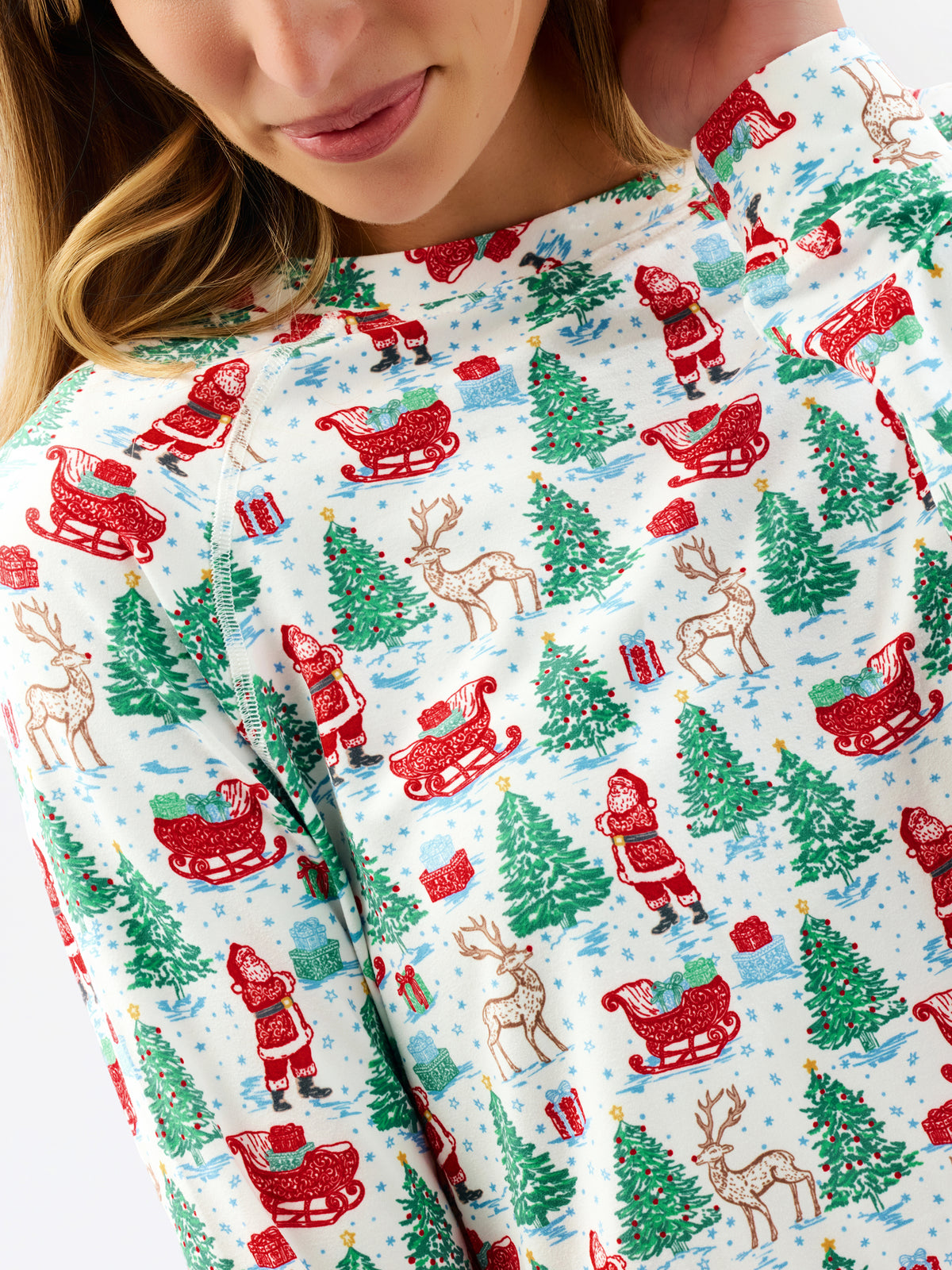 A close-up of a person wearing a white long-sleeve top with a festive Christmas print featuring Santa Claus, reindeer, sledding, and evergreen trees.