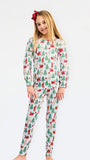 A young girl modeling a two-piece long-sleeve pajama set with a festive Christmas print of Santa Claus, elves, and Christmas trees on a white background, set against a plain light gray studio backdrop.