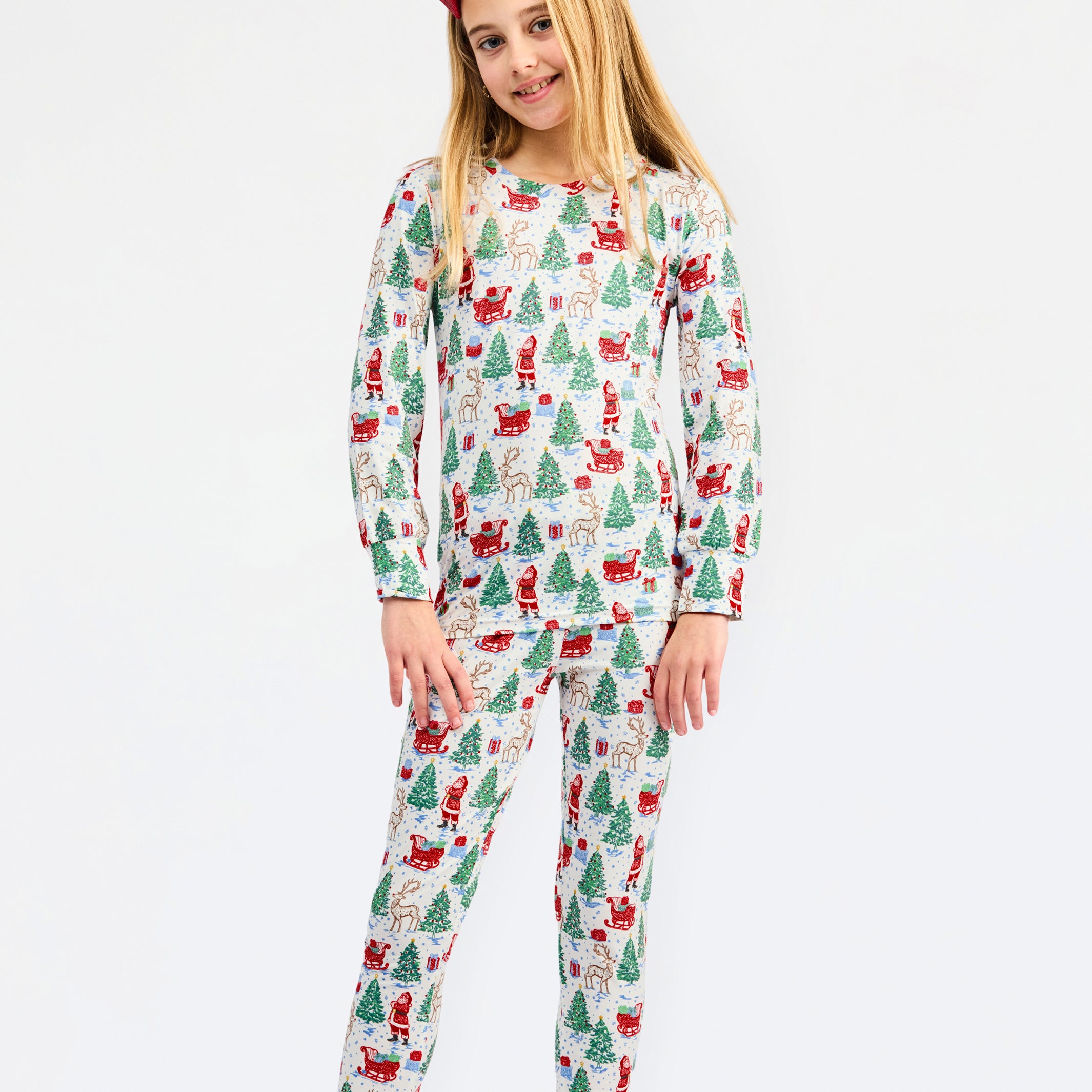 A young girl modeling a two-piece long-sleeve pajama set with a festive Christmas print of Santa Claus, elves, and Christmas trees on a white background, set against a plain light gray studio backdrop.