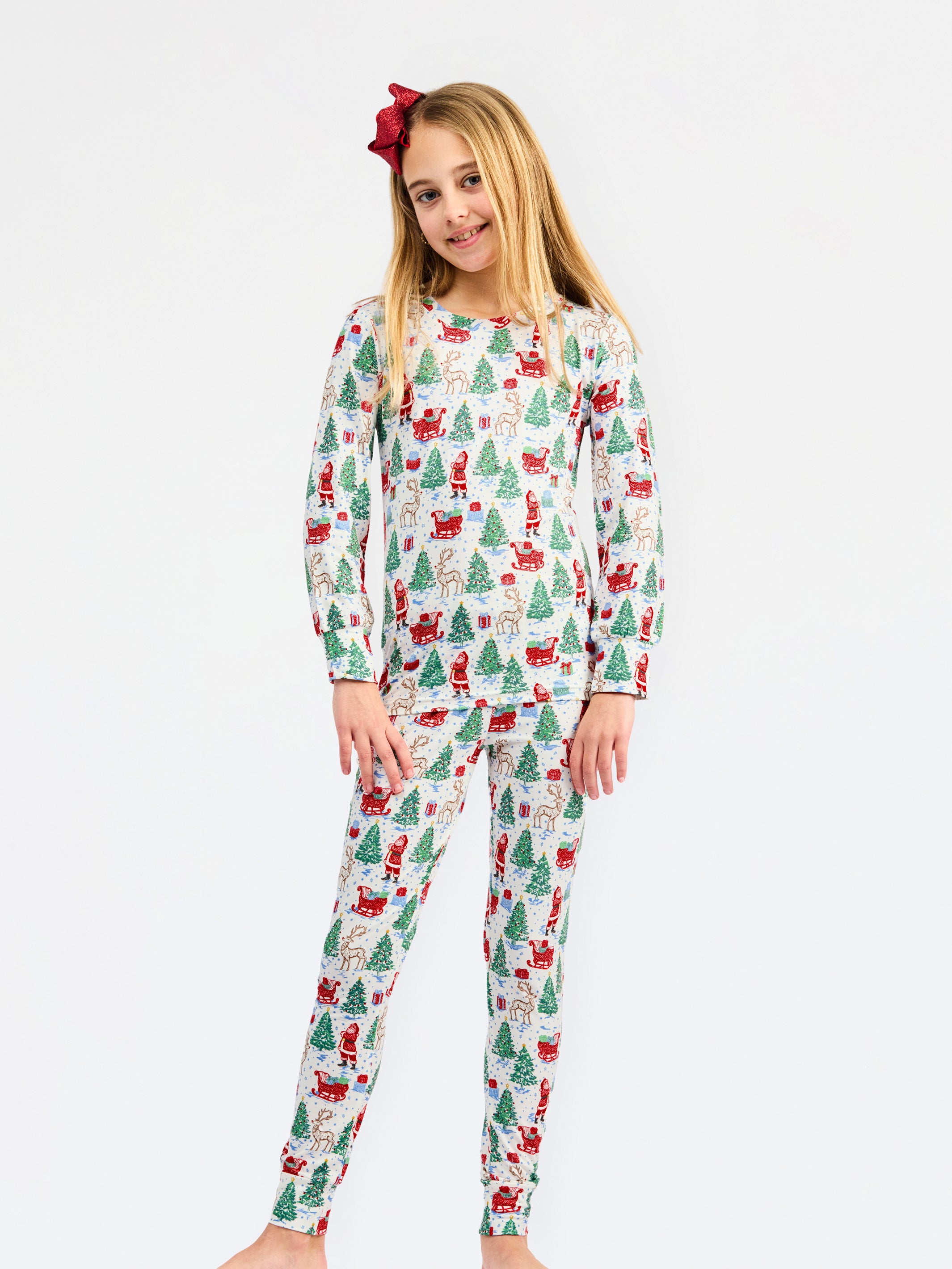 A young girl modeling a two-piece long-sleeve pajama set with a festive Christmas print of Santa Claus, elves, and Christmas trees on a white background, set against a plain light gray studio backdrop.