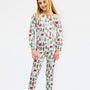 A young girl modeling a two-piece long-sleeve pajama set with a festive Christmas print of Santa Claus, elves, and Christmas trees on a white background, set against a plain light gray studio backdrop.