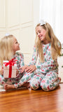 Two young girls sitting on a wooden floor in a cozy room, wearing matching Christmas-themed pajamas with a festive print. One girl holds a wrapped gift box with a red ribbon.