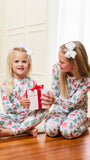 Two young girls sitting on a wooden floor wearing matching Christmas-themed pajamas with a red and green holiday print. One girl holds a white gift box with a red ribbon, and a Christmas tree is visible in the background.