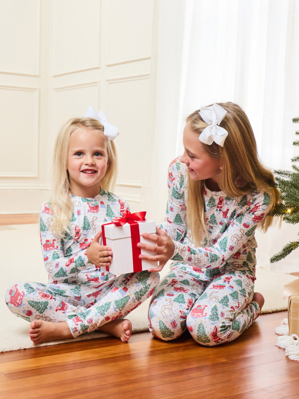 Two young girls sitting on a wooden floor wearing matching Christmas-themed pajamas with a red and green holiday print. One girl holds a white gift box with a red ribbon, and a Christmas tree is visible in the background.