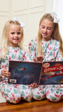Two young girls sitting on a wooden floor reading a 'Letters to Santa' book, wearing matching white Christmas-themed pajamas with a red and green holiday print.