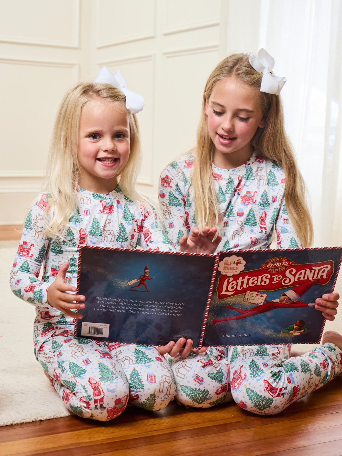 Two young girls sitting on a wooden floor reading a 'Letters to Santa' book, wearing matching white Christmas-themed pajamas with a red and green holiday print.