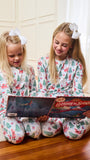Two young girls sitting on a wooden floor in a cozy room, wearing matching white Christmas-themed pajamas with a Santa Claus and tree pattern. They are looking at a 'Letters to Santa' box together.