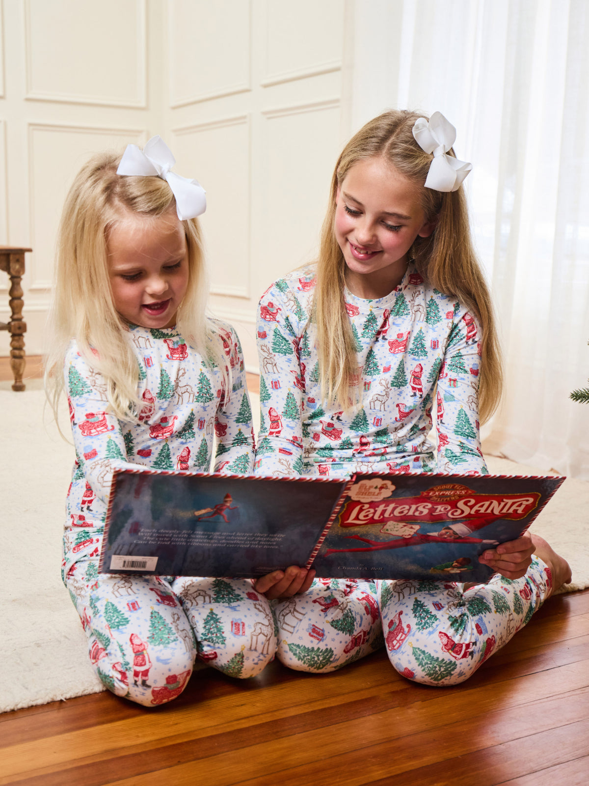 Two young girls sitting on a wooden floor in a cozy room, wearing matching white Christmas-themed pajamas with a Santa Claus and tree pattern. They are looking at a 'Letters to Santa' box together.