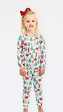 A young girl with blonde hair and a red bow stands against a white studio background wearing a long-sleeve pajama set with a festive Christmas print featuring Santa Claus, reindeer, and green trees.