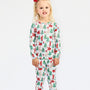 A young girl with blonde hair and a red bow stands against a white studio background wearing a long-sleeve pajama set with a festive Christmas print featuring Santa Claus, reindeer, and green trees.