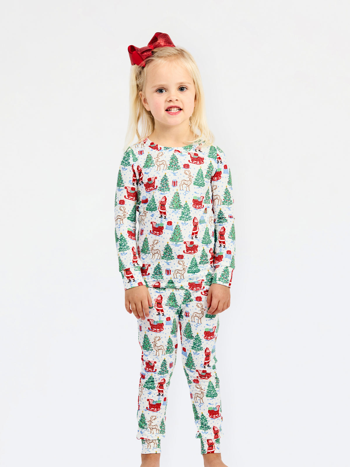 A young girl with blonde hair and a red bow stands against a white studio background wearing a long-sleeve pajama set with a festive Christmas print featuring Santa Claus, reindeer, and green trees.