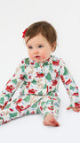 A baby sitting on a white studio background wearing a long-sleeve holiday-themed pajama romper with a festive print of Santa Claus, reindeer, Christmas trees, and sleighs. The baby has a small red bow in their hair.