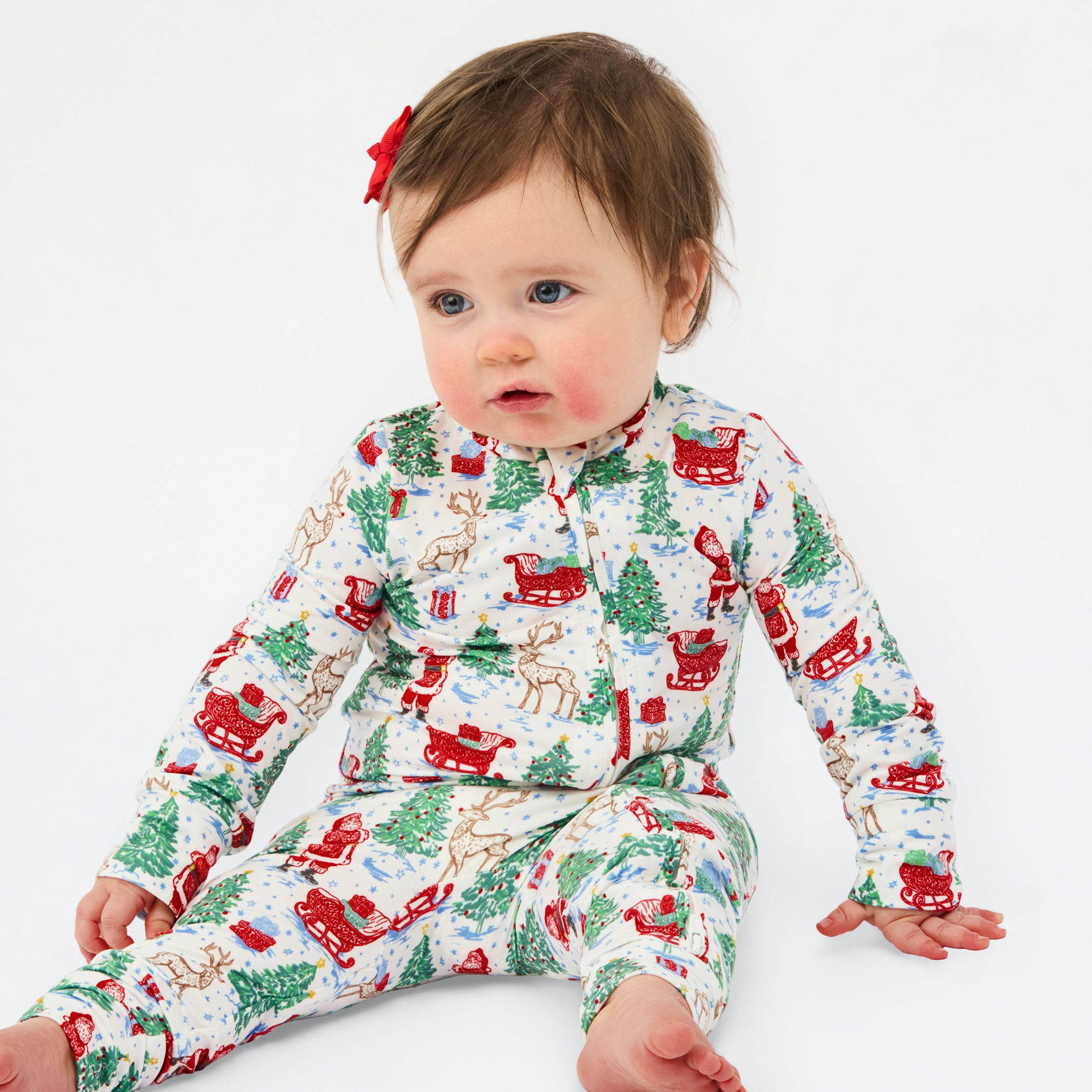 A baby sitting on a white studio background wearing a long-sleeve holiday-themed pajama romper with a festive print of Santa Claus, reindeer, Christmas trees, and sleighs. The baby has a small red bow in their hair.
