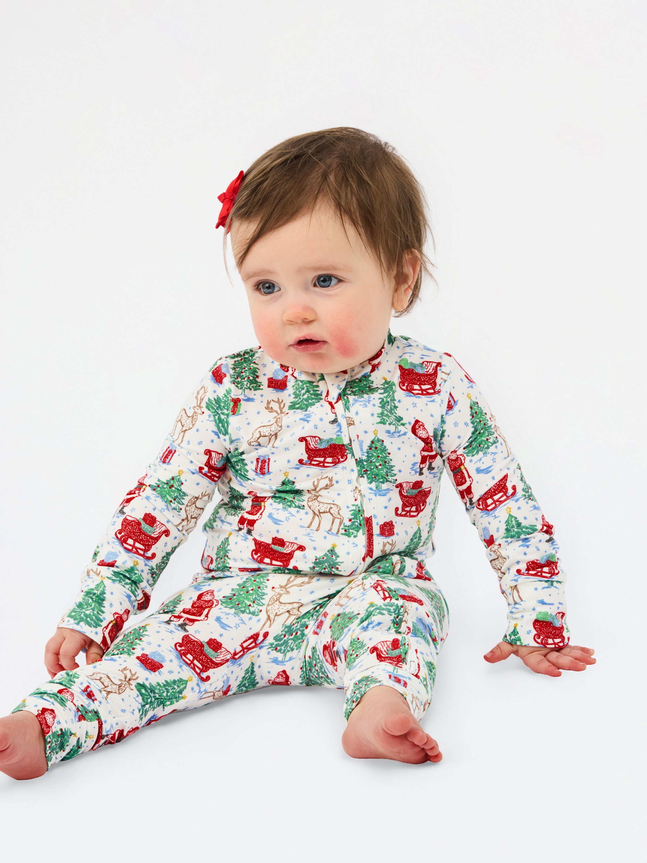 A baby sitting on a white studio background wearing a long-sleeve holiday-themed pajama romper with a festive print of Santa Claus, reindeer, Christmas trees, and sleighs. The baby has a small red bow in their hair.