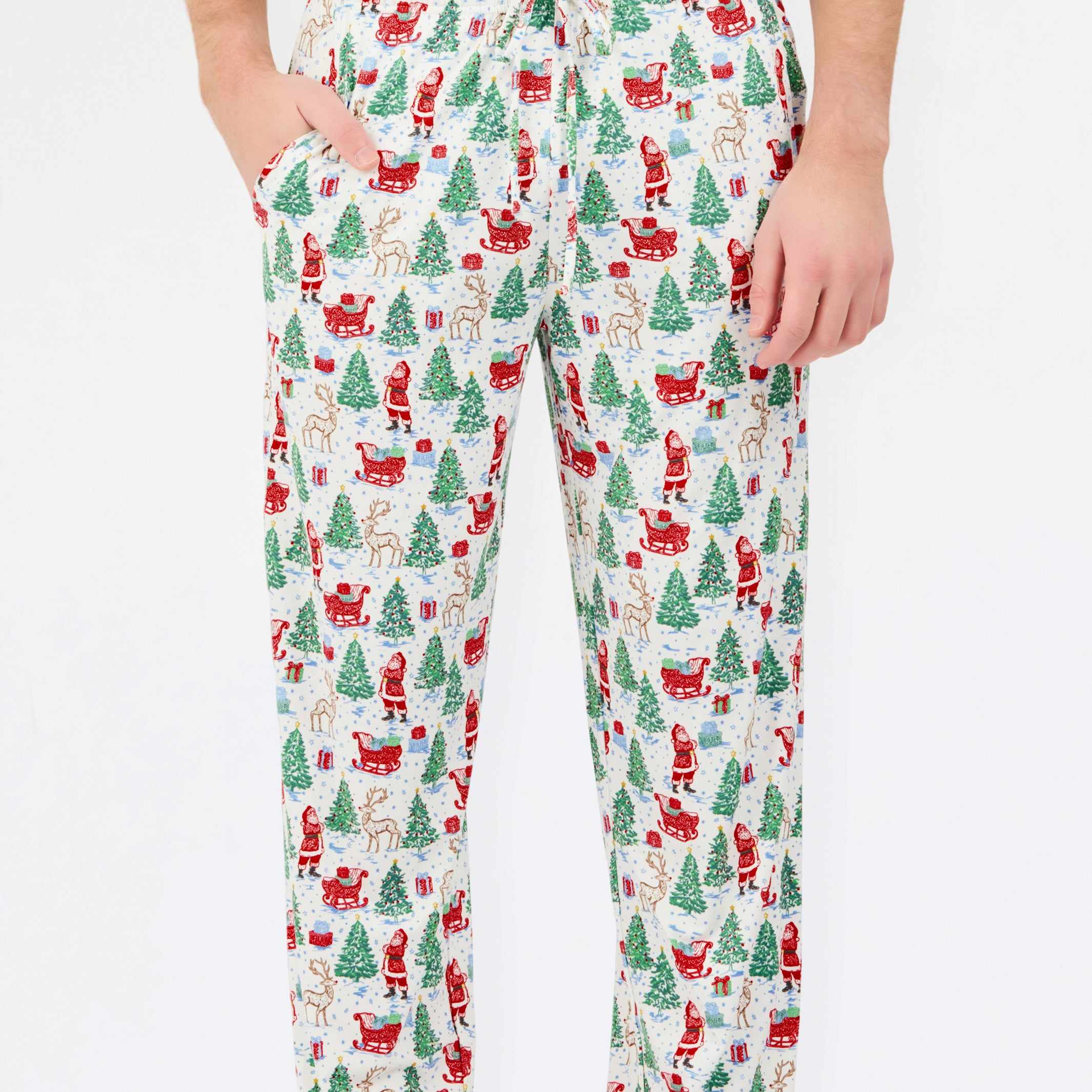 A person wearing holiday-themed lounge pants with an all-over Christmas print featuring Santa Claus, reindeer, trees, and sleighs on a white background.