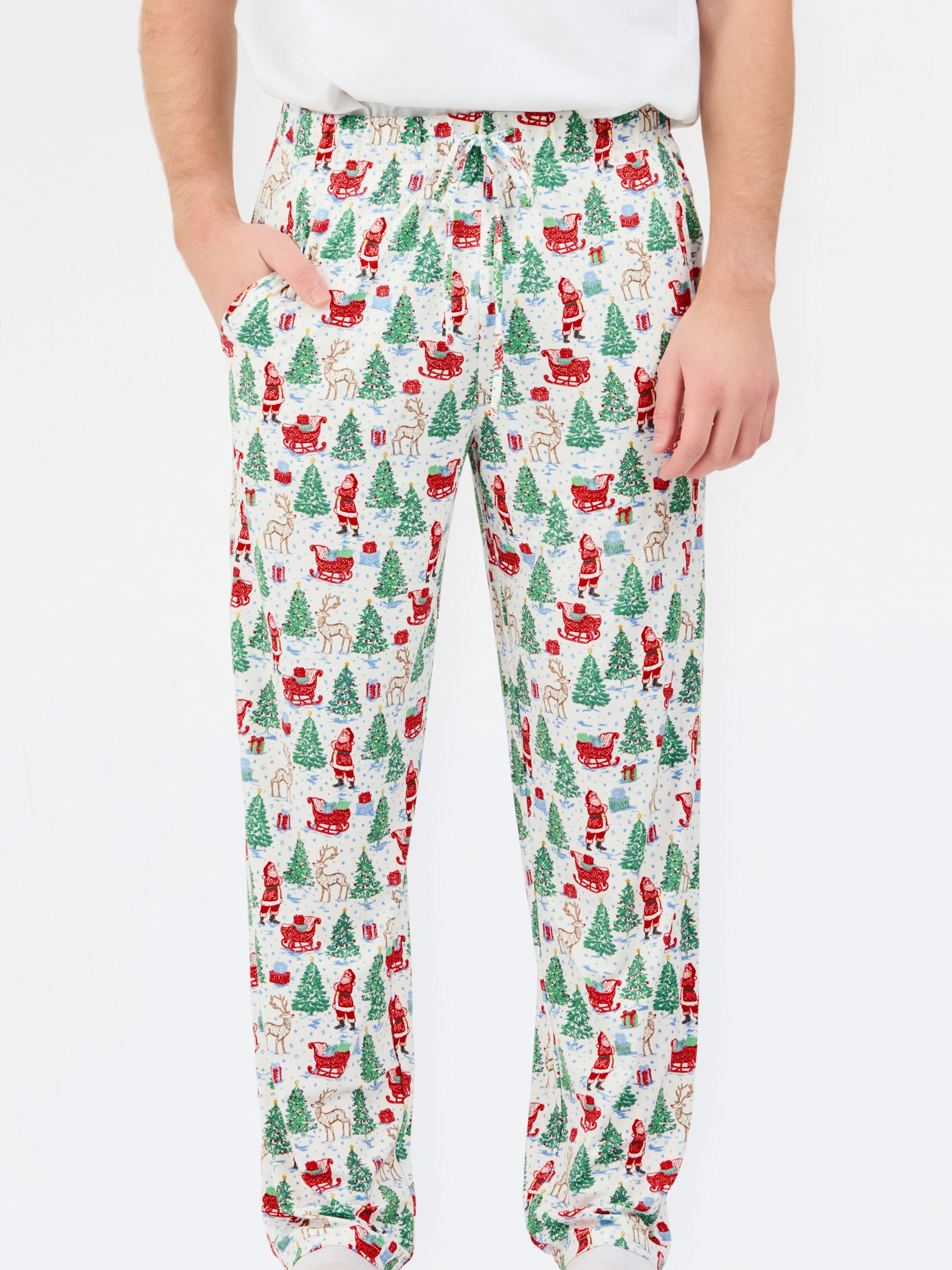 A person wearing holiday-themed lounge pants with an all-over Christmas print featuring Santa Claus, reindeer, trees, and sleighs on a white background.