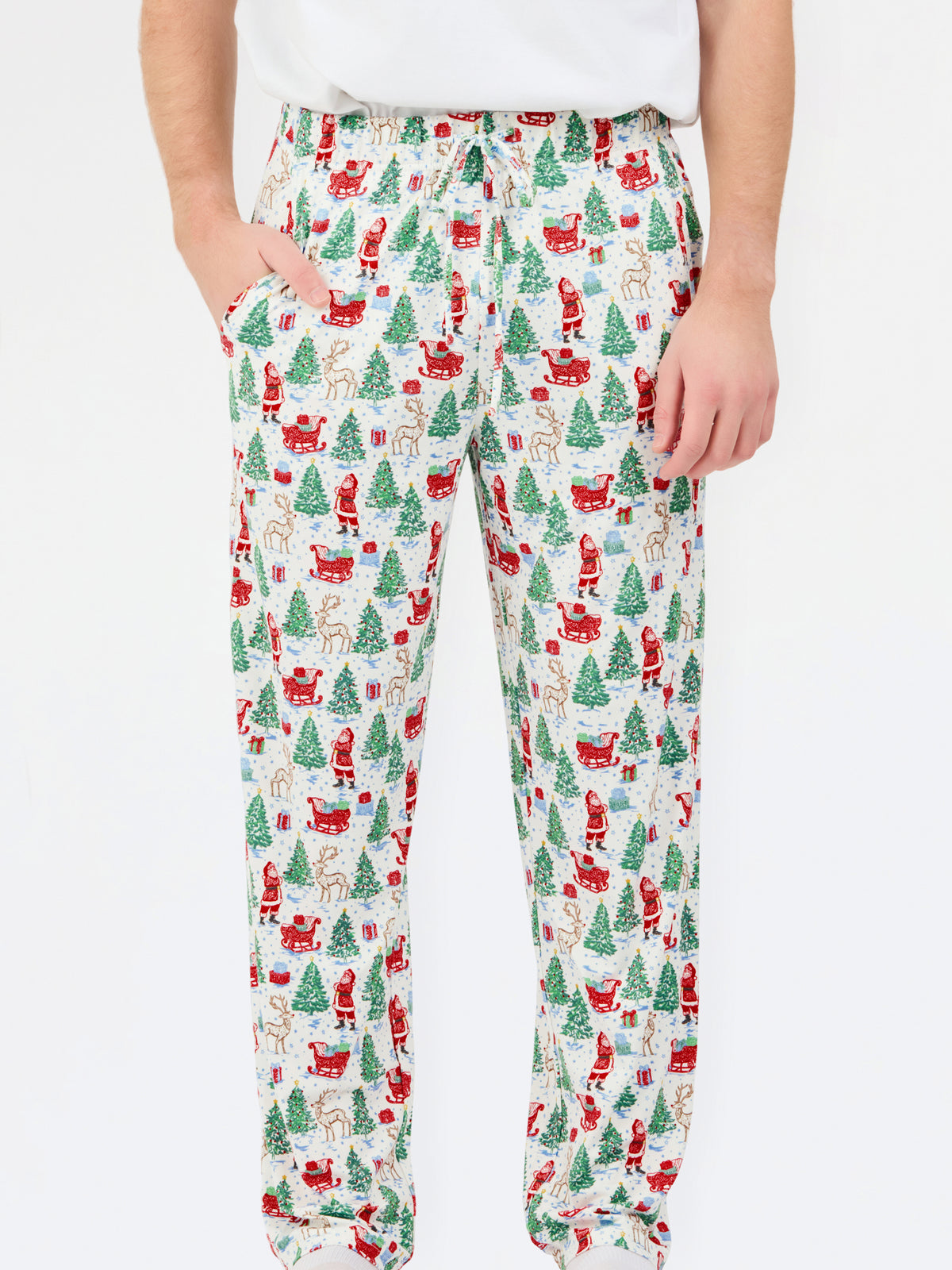 A person wearing holiday-themed lounge pants with an all-over Christmas print featuring Santa Claus, reindeer, trees, and sleighs on a white background.