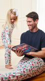 A man and a young girl wearing matching Christmas-themed pajamas sit and stand together in a warmly lit room, with the man holding a book. The pajamas have a white base with a festive print of red Santa Claus figures and green Christmas trees.
