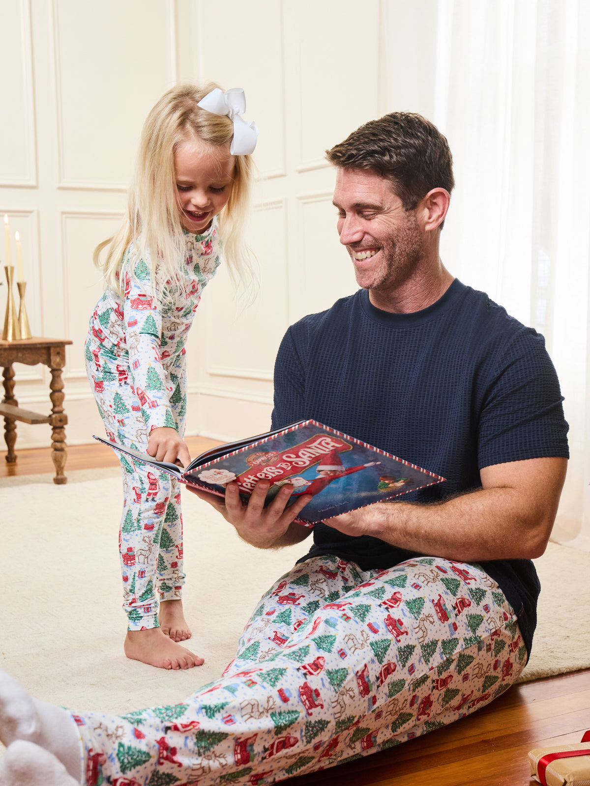 A man and a young girl wearing matching Christmas-themed pajamas sit and stand together in a warmly lit room, with the man holding a book. The pajamas have a white base with a festive print of red Santa Claus figures and green Christmas trees.