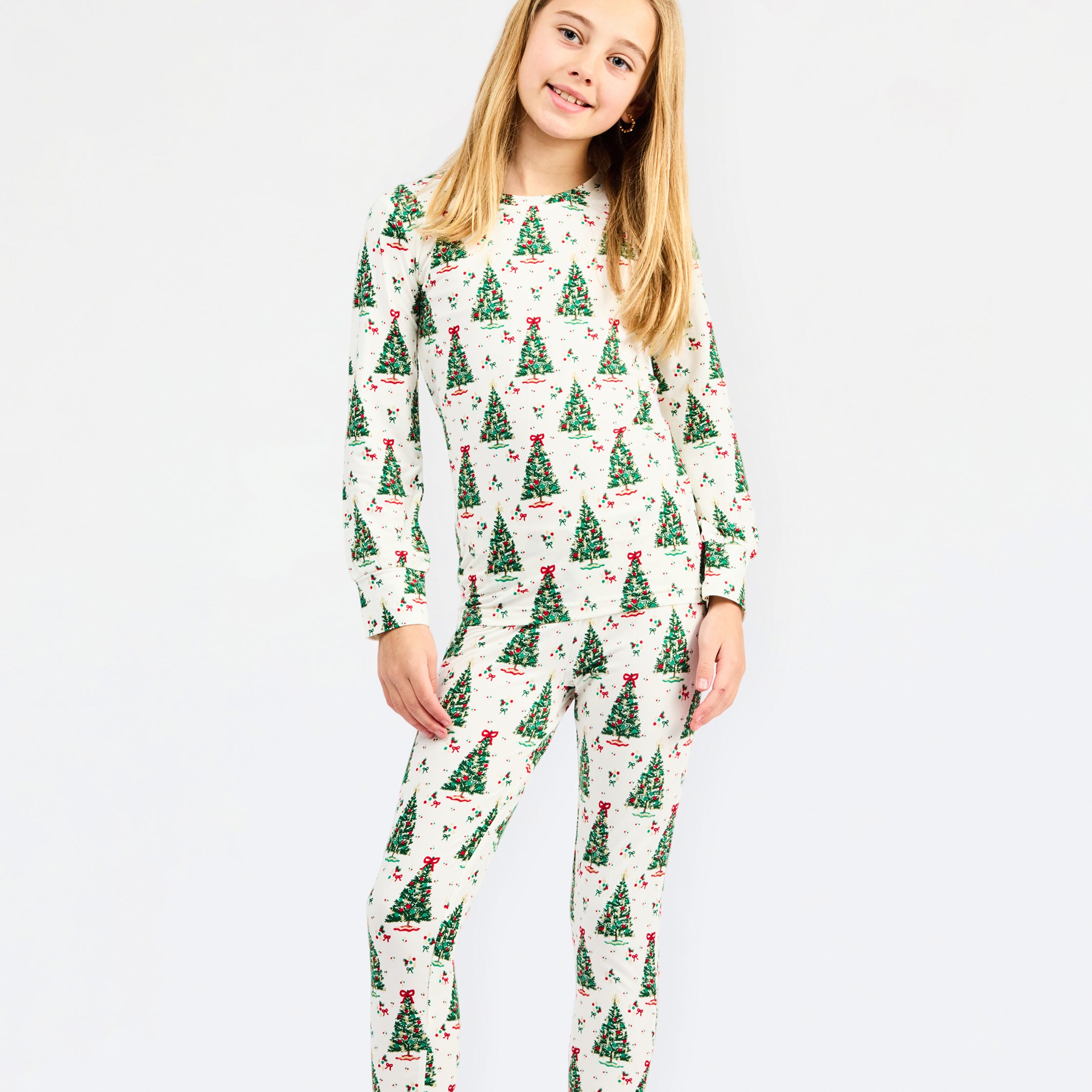 A young girl with blonde hair and a red bow headband is wearing a two-piece long-sleeve pajama set with a Christmas tree pattern, standing against a plain white background.