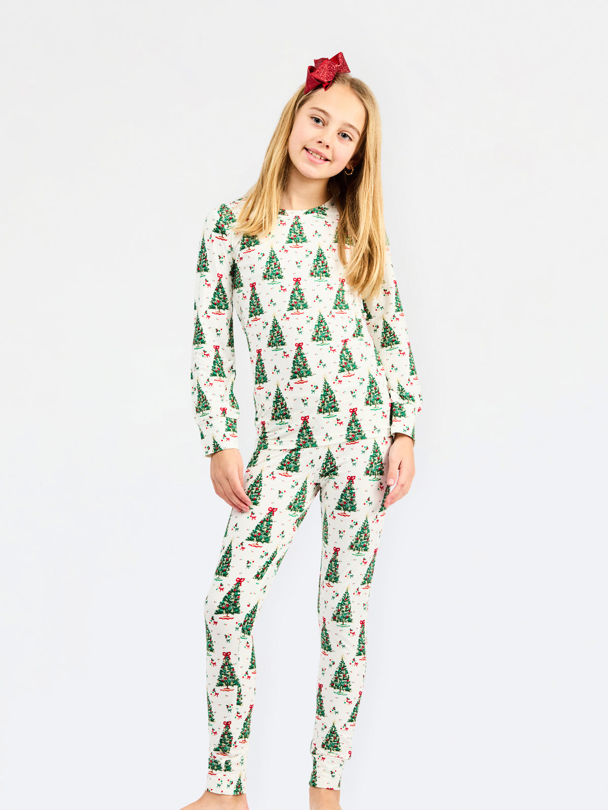 A young girl with blonde hair and a red bow headband is wearing a two-piece long-sleeve pajama set with a Christmas tree pattern, standing against a plain white background.