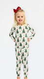 A young girl with blonde hair and a red bow stands against a white background wearing a long-sleeve pajama set with a green Christmas tree pattern on a cream base.