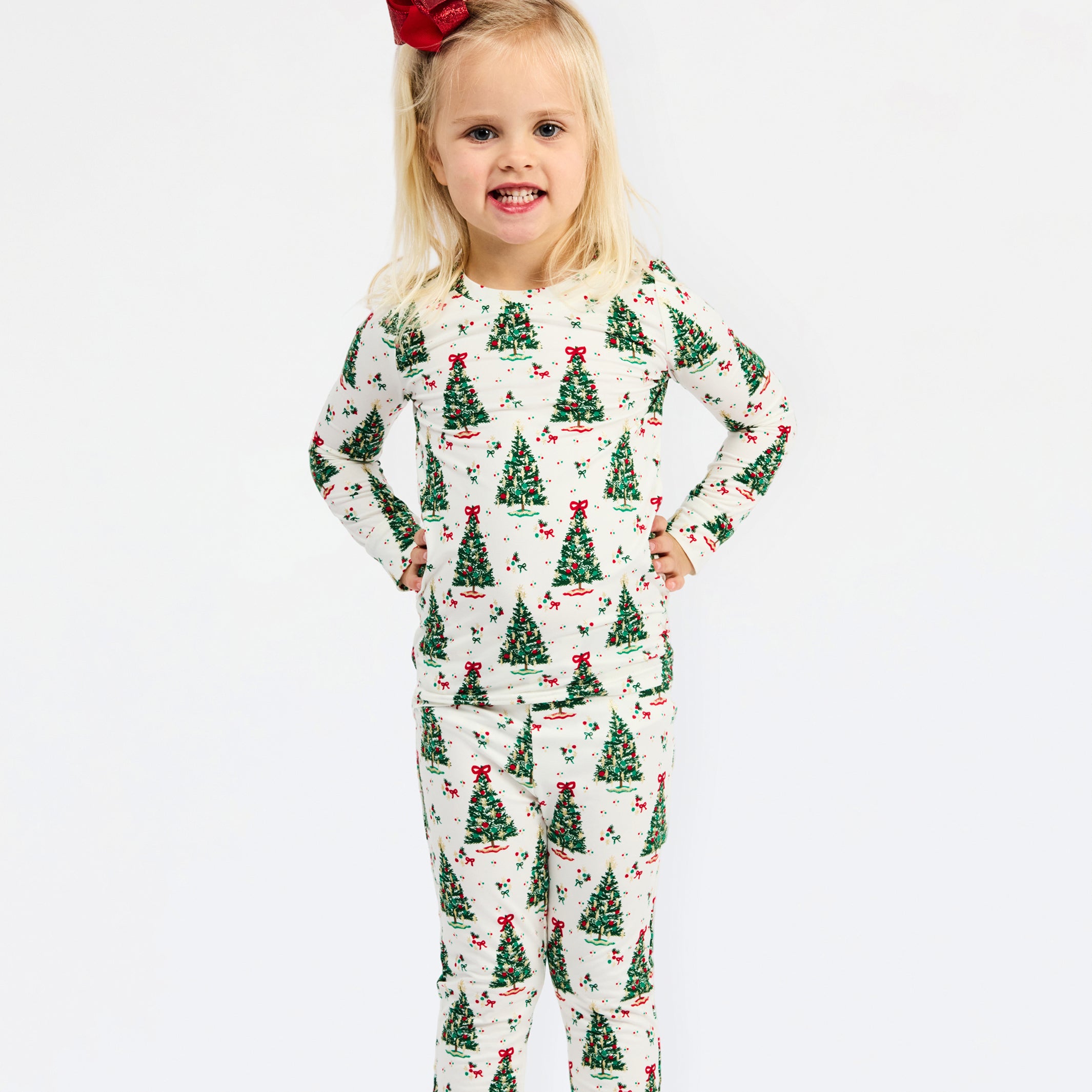 A young girl with blonde hair and a red bow stands against a white background wearing a long-sleeve pajama set with a green Christmas tree pattern on a cream base.