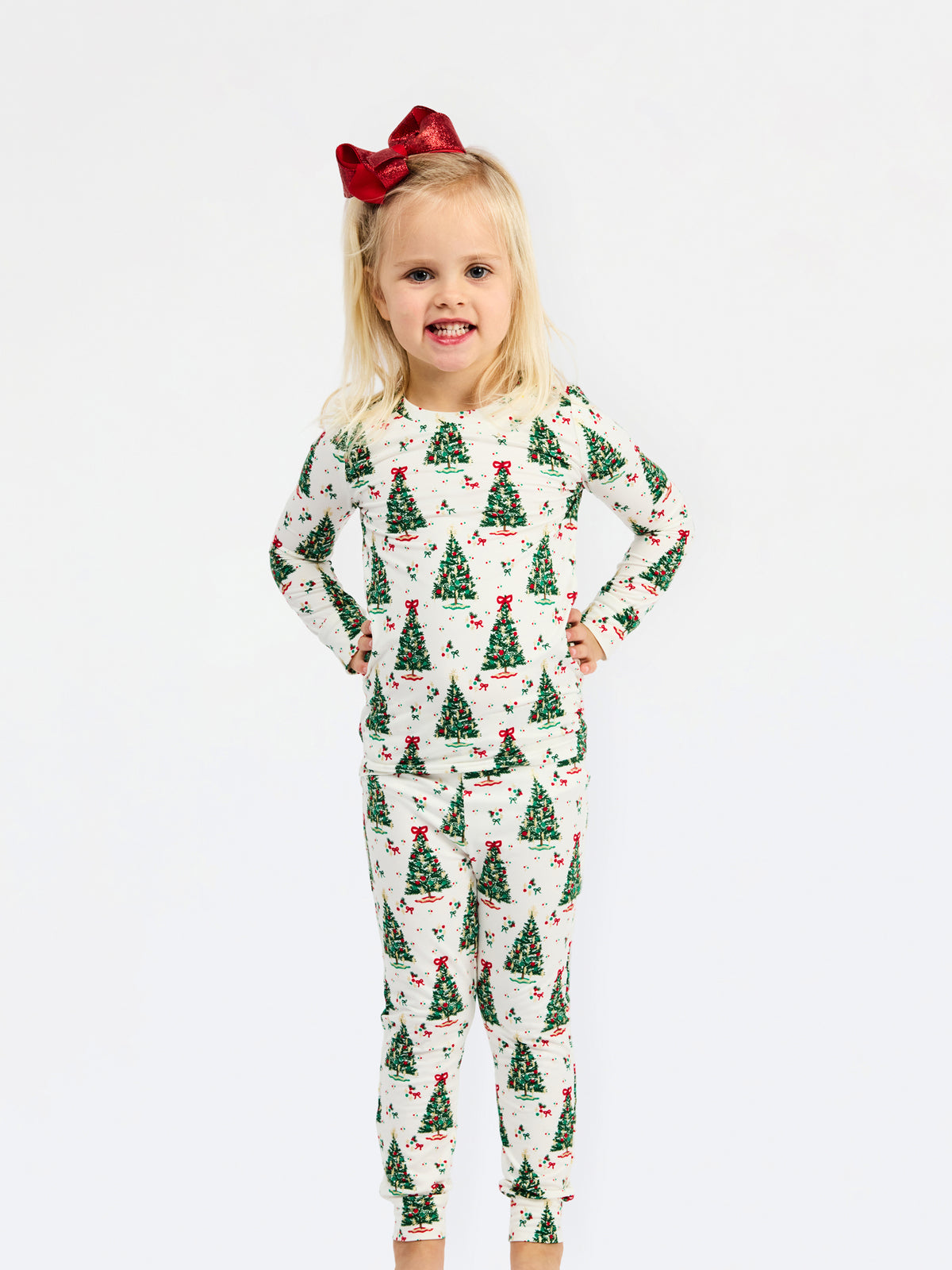 A young girl with blonde hair and a red bow stands against a white background wearing a long-sleeve pajama set with a green Christmas tree pattern on a cream base.