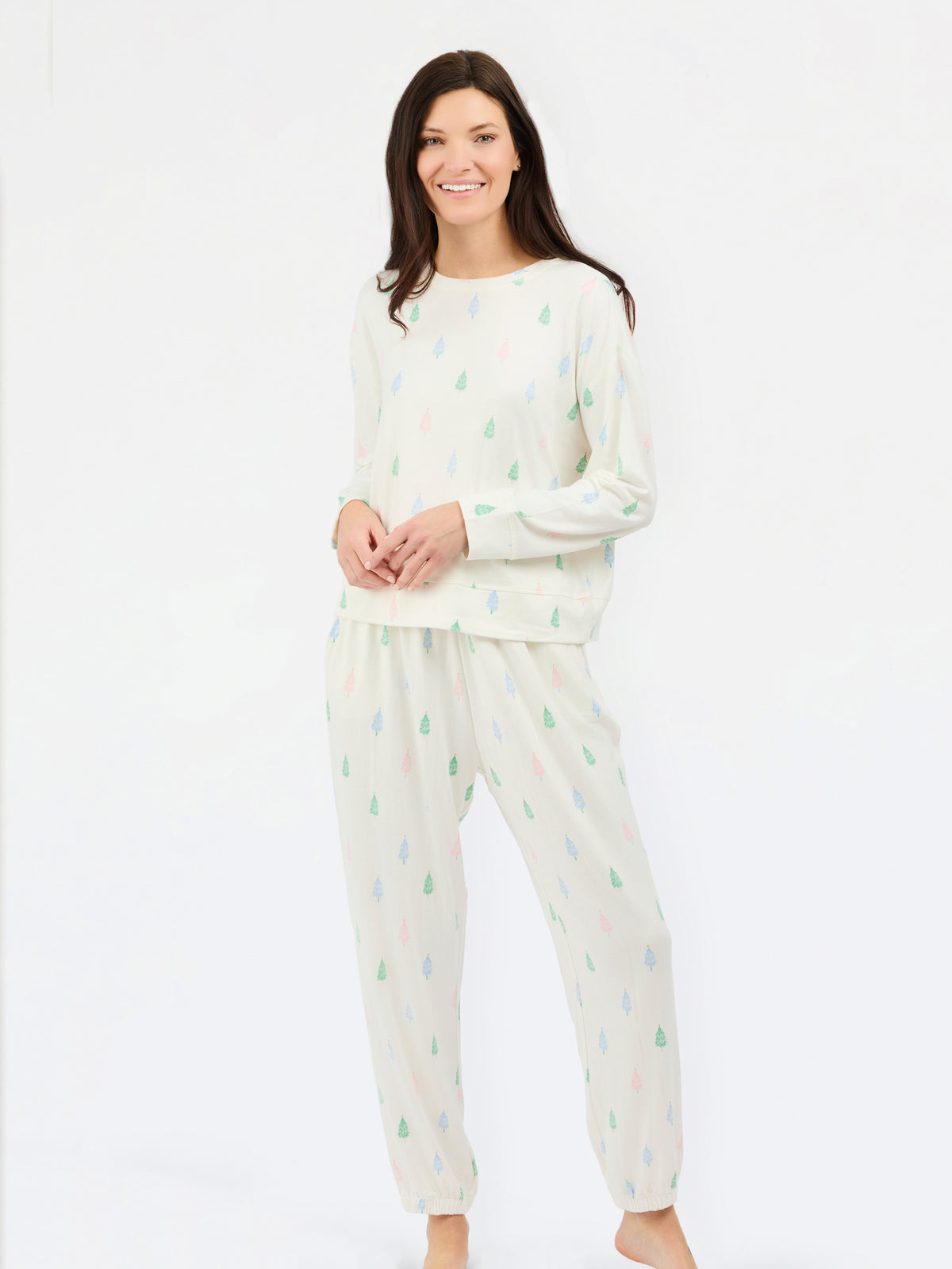 A woman modeling a two-piece pajama set consisting of a long-sleeve top and matching pants, both in an off-white color with a small, scattered pine tree pattern in light green, blue, and pink.
