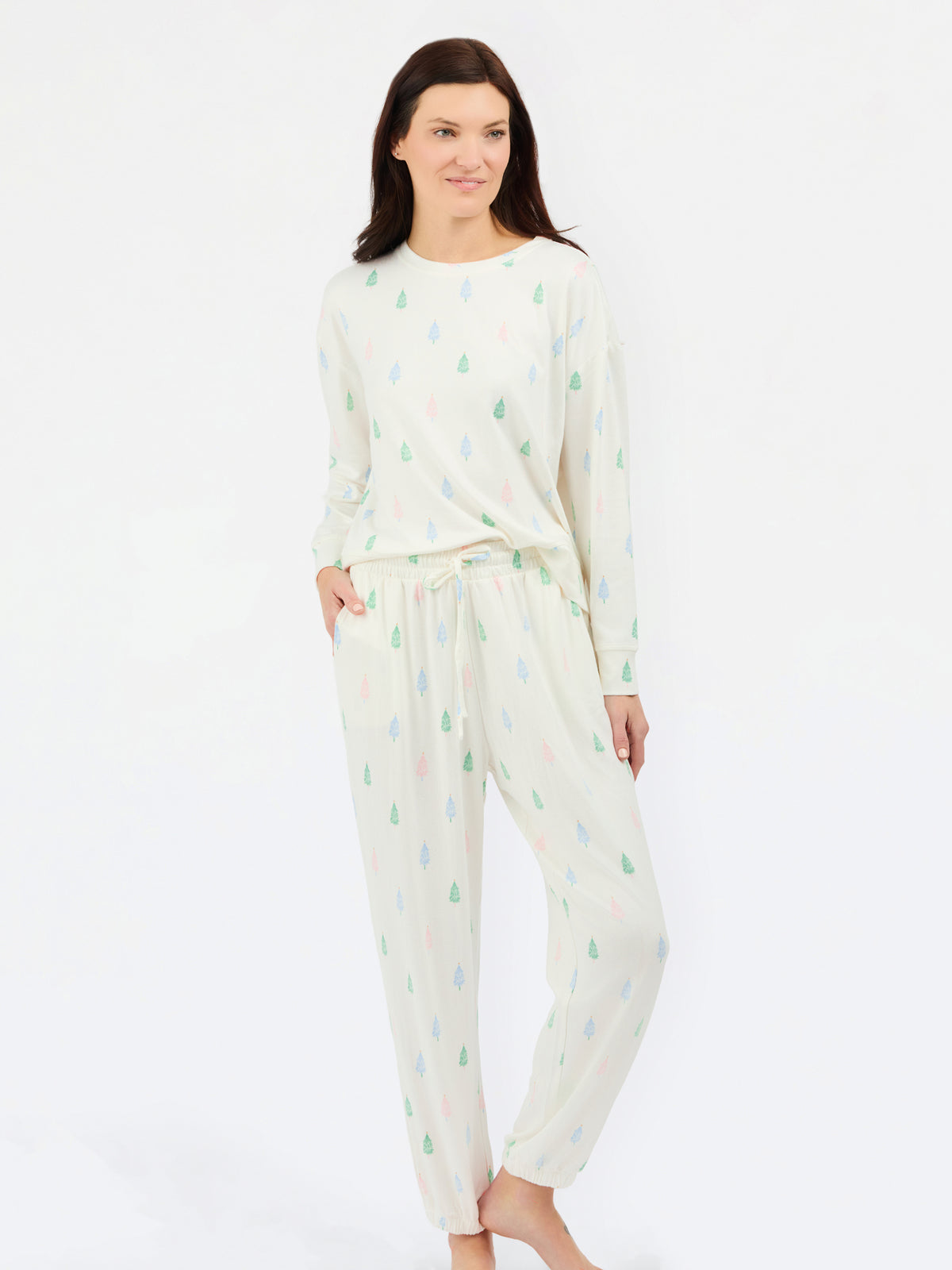 A woman modeling a two-piece long-sleeve pajama set with a white base and a small repeating pattern of green, blue, and pink pine trees, photographed against a plain white studio background.