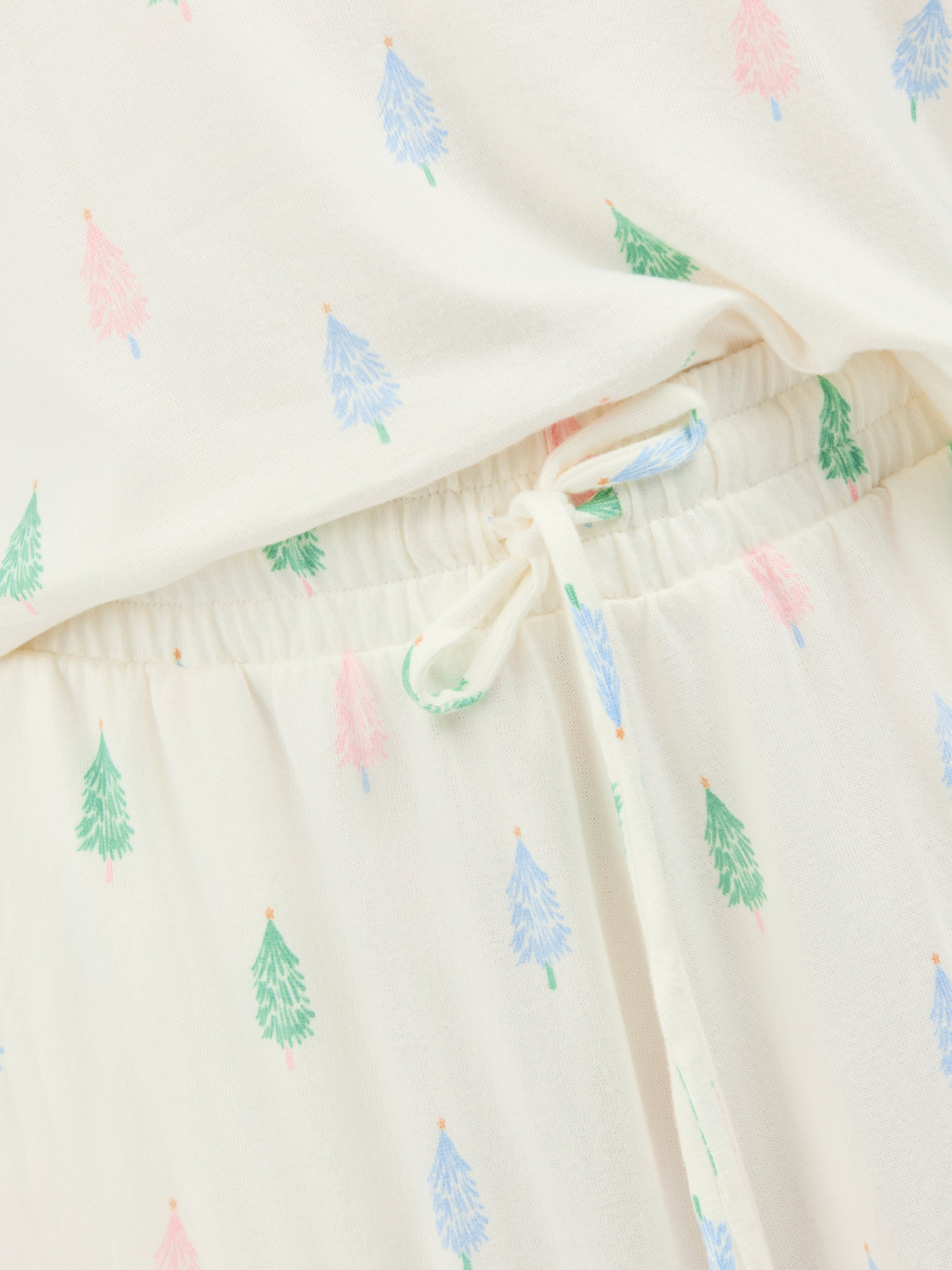 Close-up of a cream-colored pajama set with a repeating pattern of small pine trees in green, blue, and pink, featuring an elastic waistband and drawstring.
