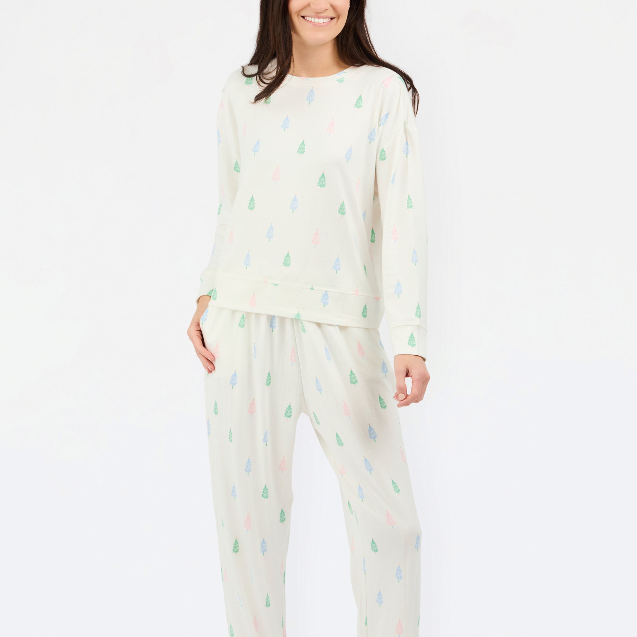 A woman modeling a two-piece pajama set featuring a cream-colored long-sleeve top and matching pants, both decorated with a small, repeating multicolor pine tree pattern in green, blue, and pink.