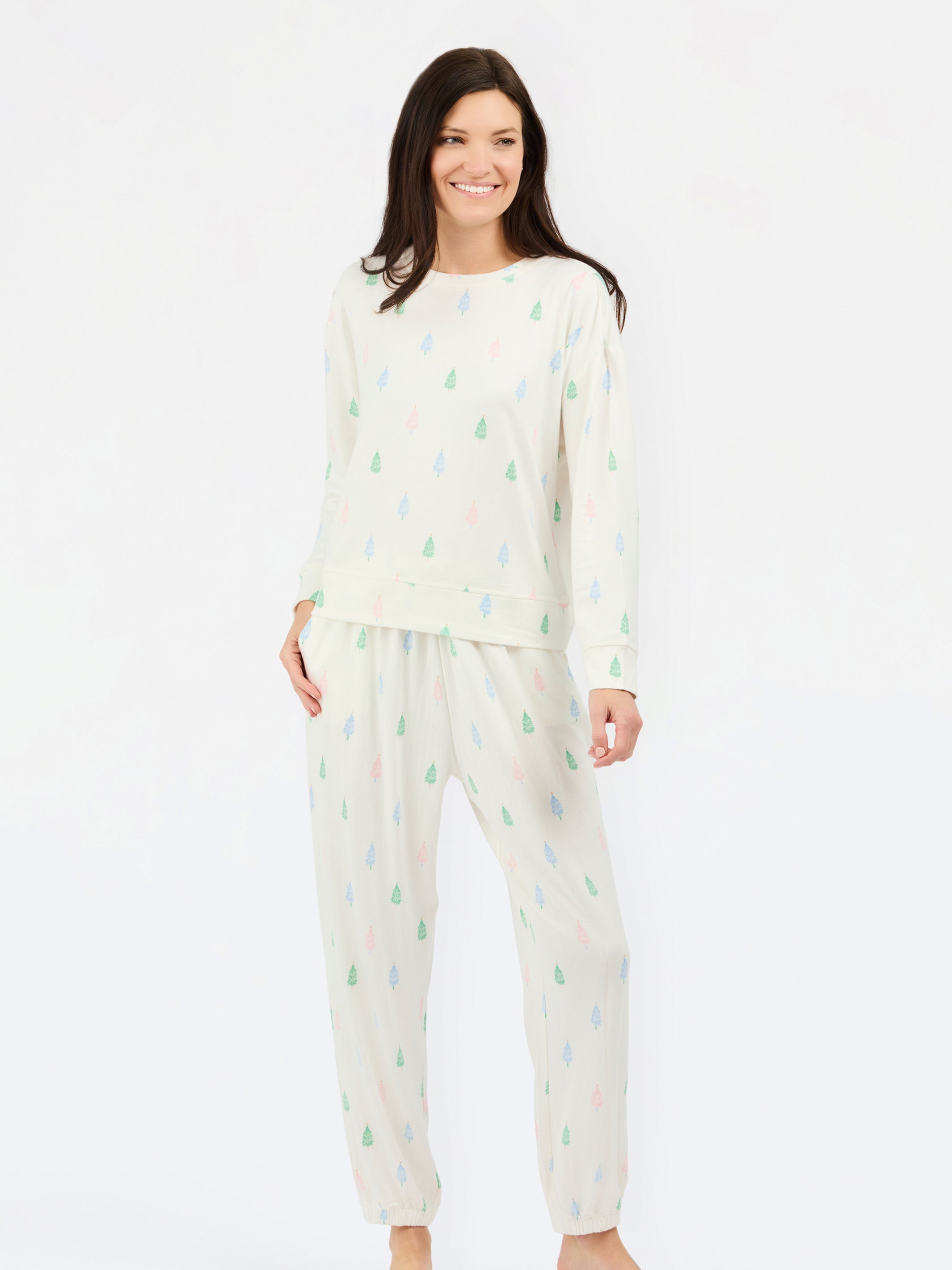 A woman modeling a two-piece pajama set featuring a cream-colored long-sleeve top and matching pants, both decorated with a small, repeating multicolor pine tree pattern in green, blue, and pink.