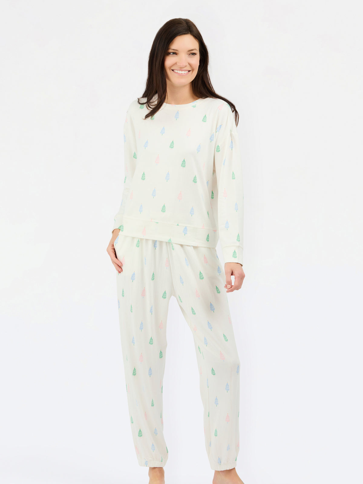 A woman modeling a two-piece pajama set featuring a cream-colored long-sleeve top and matching pants, both decorated with a small, repeating multicolor pine tree pattern in green, blue, and pink.