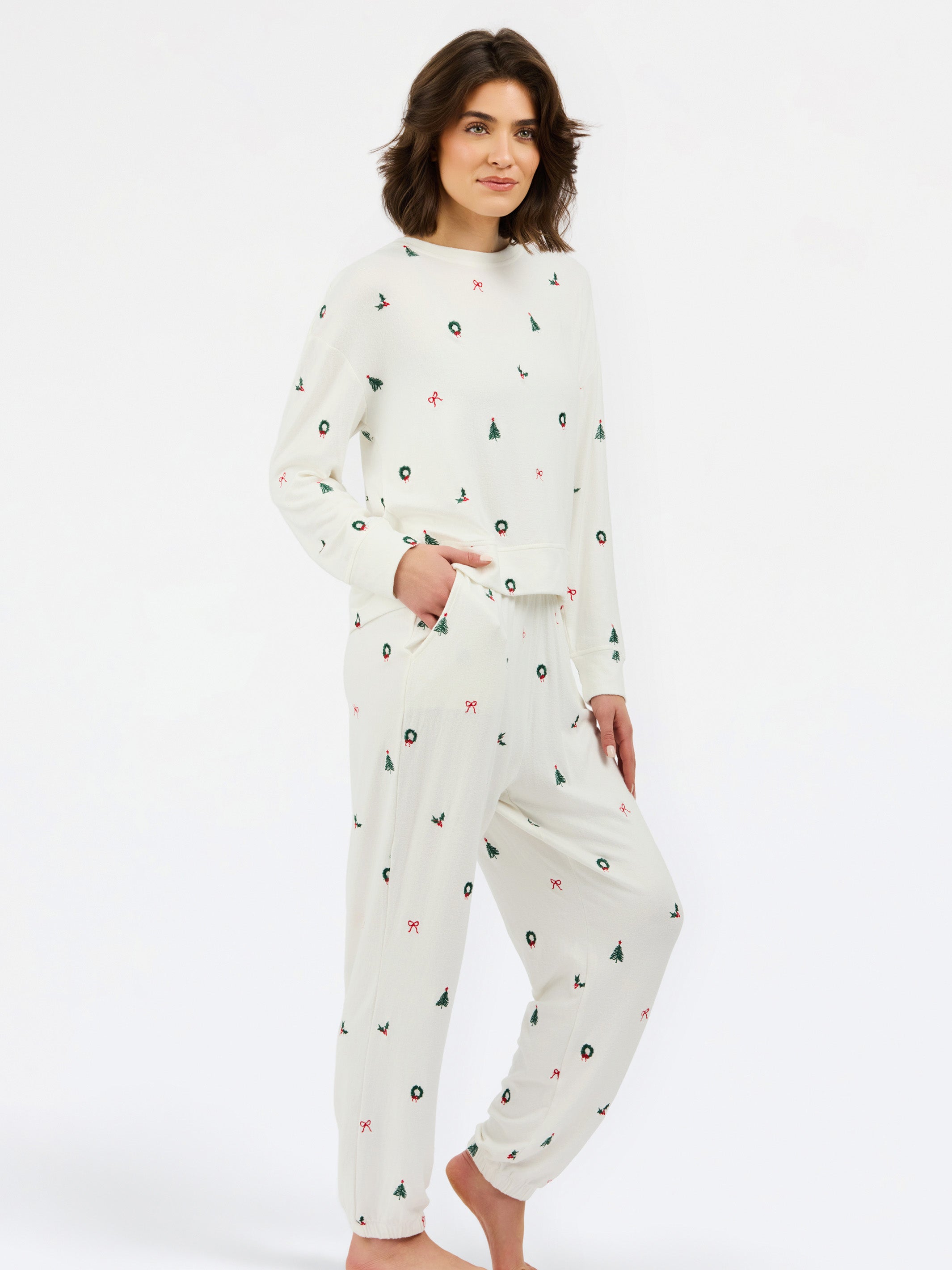 A woman modeling a matching two-piece pajama set with a long-sleeve top and jogger pants, featuring a white base with a small scattered print of black circles, green trees, and red stars, against a plain white studio background.