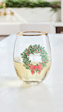A clear stemless wine glass with a gold rim, featuring a printed green wreath with red berries and a red bow, sitting on a light countertop with a blurred kitchen background.
