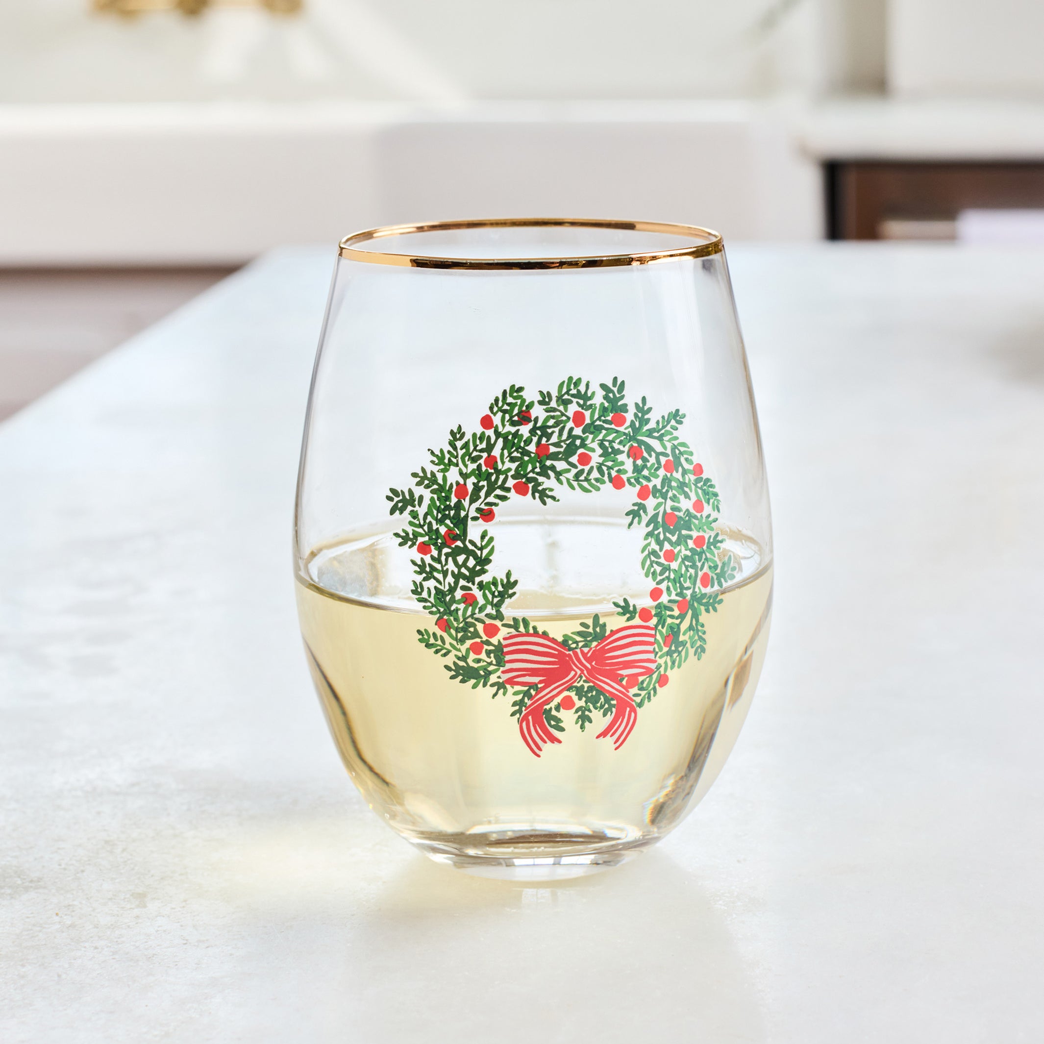 A clear stemless wine glass with a gold rim, featuring a printed green wreath with red berries and a red bow, sitting on a light countertop with a blurred kitchen background.