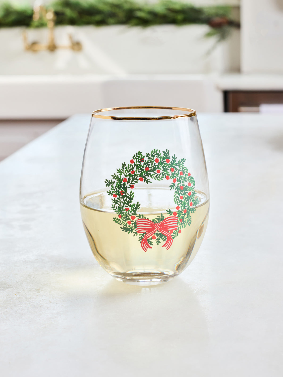 A clear stemless wine glass with a gold rim, featuring a printed green wreath with red berries and a red bow, sitting on a light countertop with a blurred kitchen background.