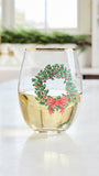 A clear stemless wine glass with a gold rim, filled with white wine, featuring a green holly wreath and red bow graphic on the side, set on a white countertop in a bright kitchen.
