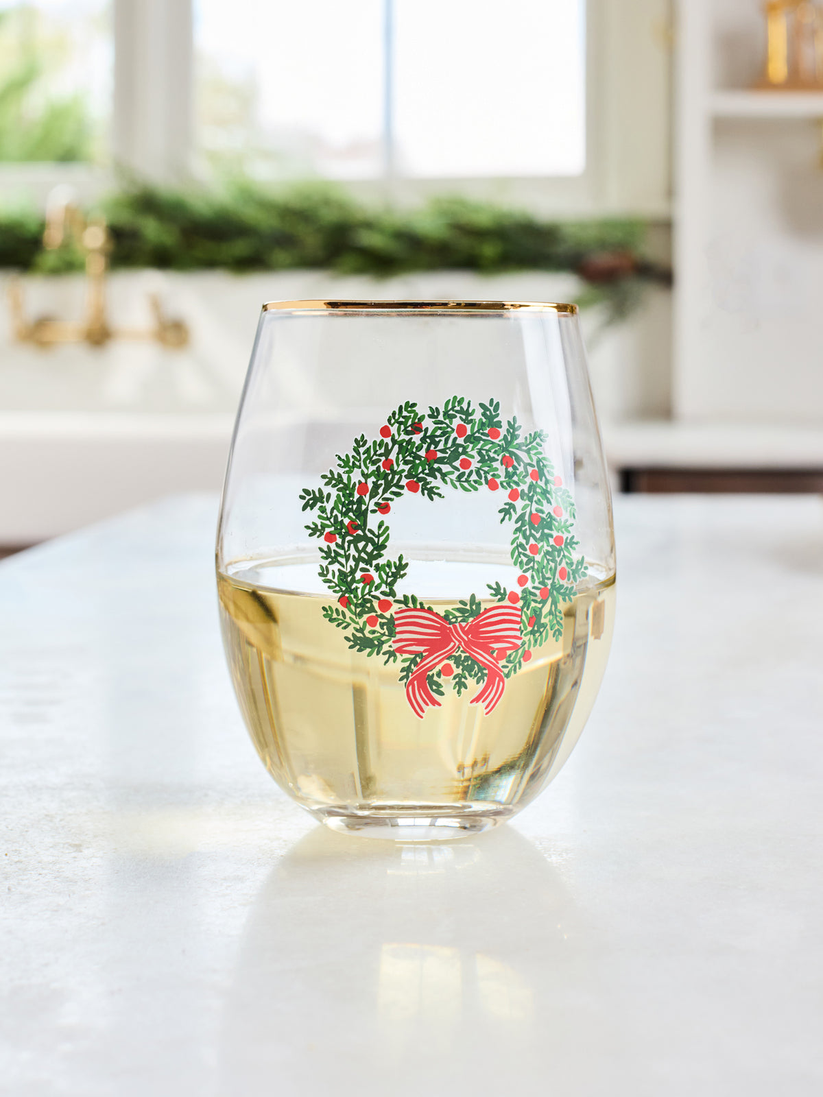 A clear stemless wine glass with a gold rim, filled with white wine, featuring a green holly wreath and red bow graphic on the side, set on a white countertop in a bright kitchen.