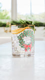 A clear glass tumbler with a gold rim and a festive green wreath and red bow print, filled with a clear cocktail and garnished with a mint leaf and orange slice, set on a light countertop with a blurred kitchen background.