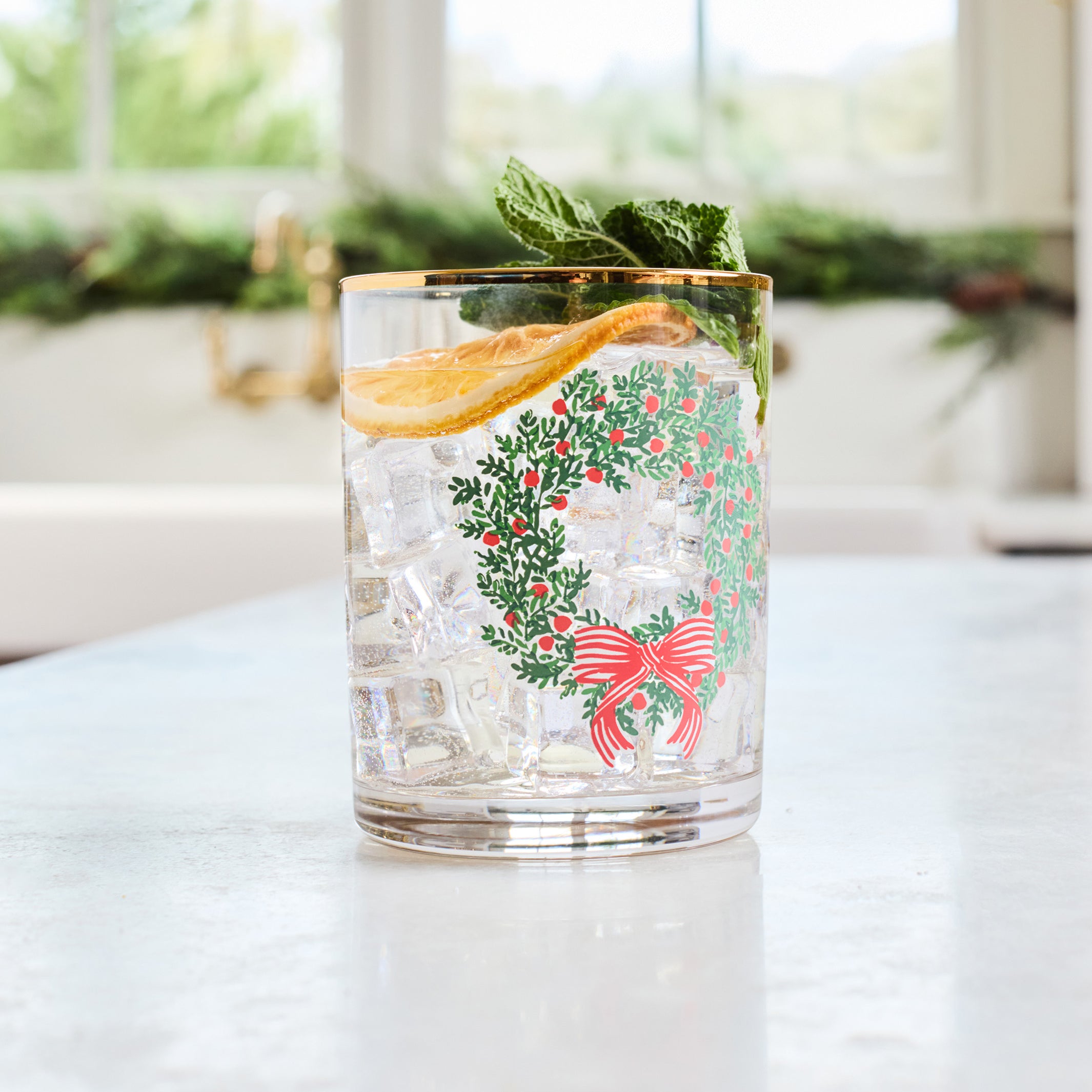 A clear glass tumbler with a gold rim and a festive green wreath and red bow print, filled with a clear cocktail and garnished with a mint leaf and orange slice, set on a light countertop with a blurred kitchen background.